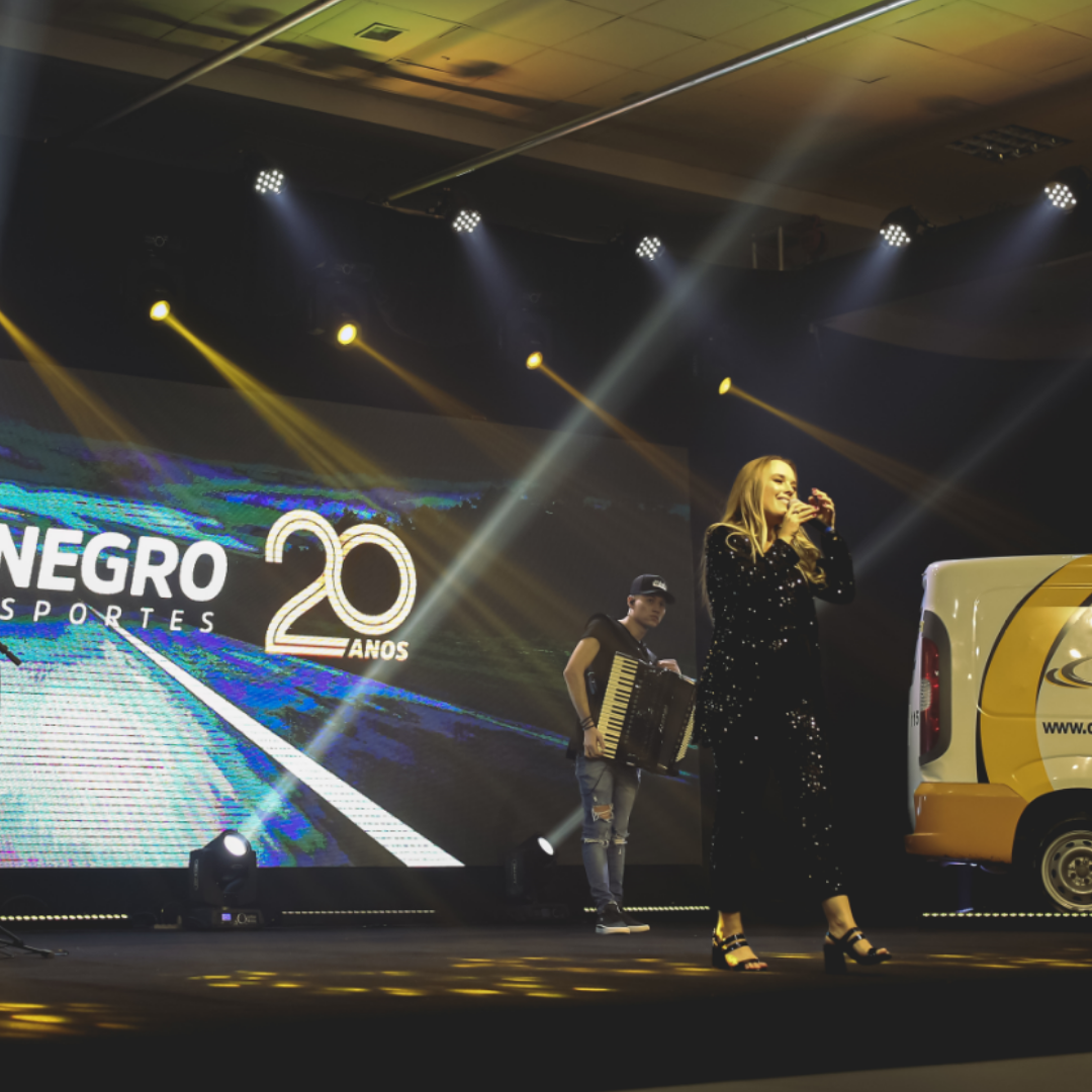 Mulher loira, vestida de preto, no palco apresentando uma live show.