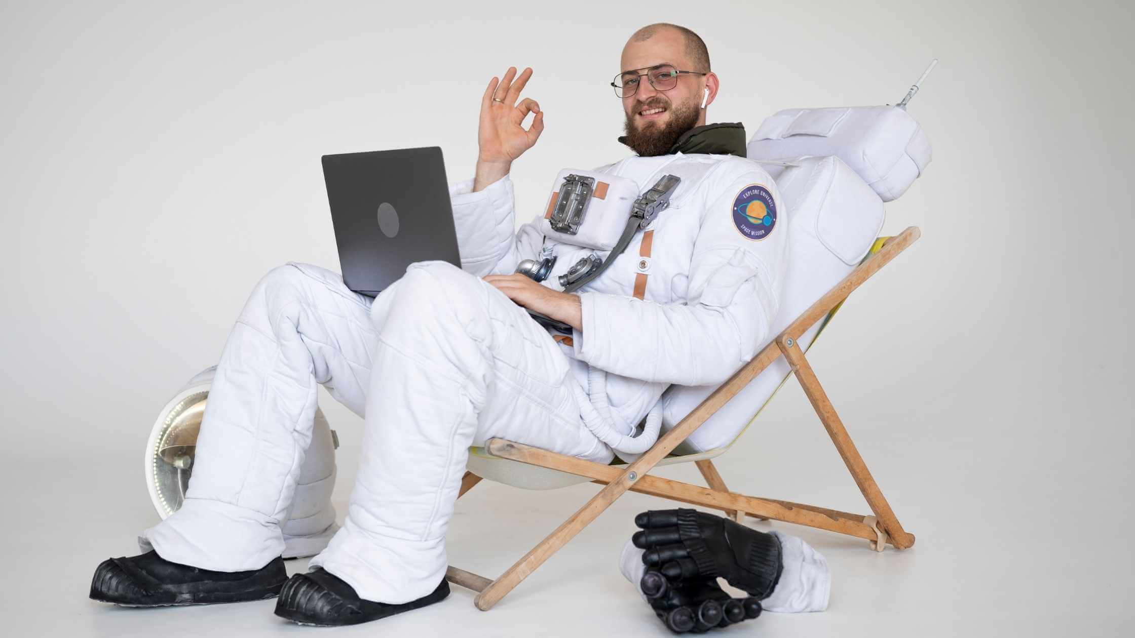 Homem branco, cabelo raspado, barba ruiva, vestido de astronauta, sentado em cadeira espreguiçadeira branca. Ele usa um laptop no colo e faz o sinal de OK com os dedos.